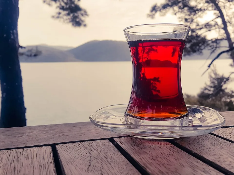 Ahşap masada tabağıyla ince belli bardakta ışıkta parlayan demli çay, arkada su ve tepeler